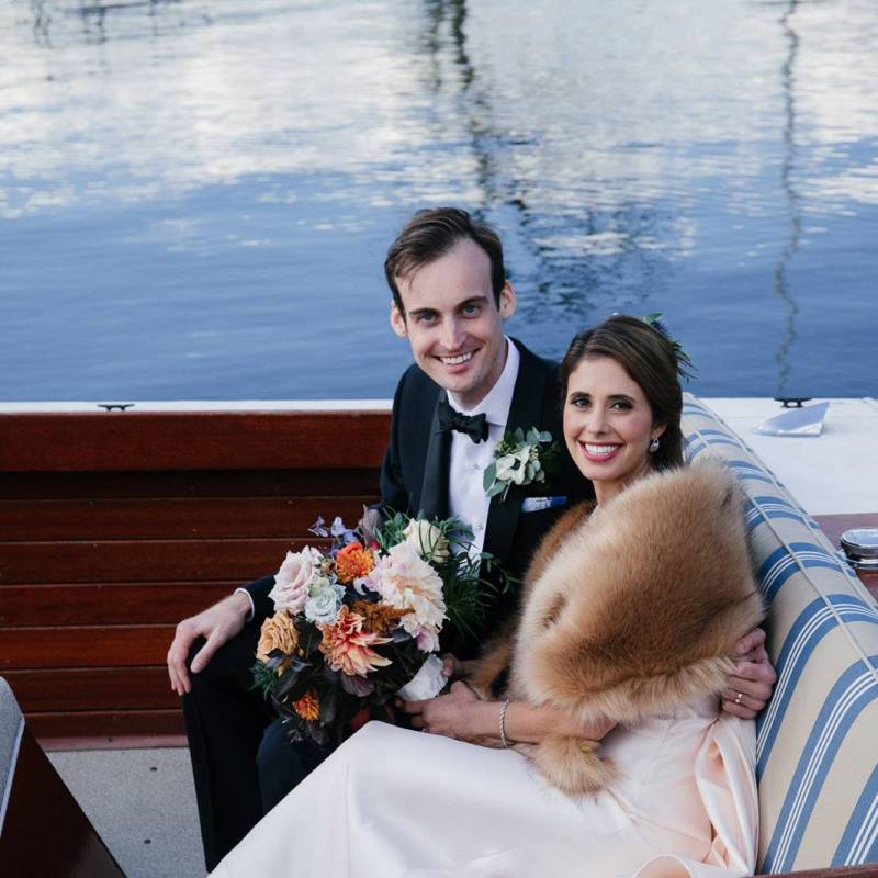 A bride wearing a fur shawl sitting next to her new husband in a tux in a boat on their wedding day in Vermont